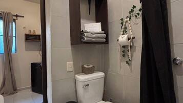 A bathroom with a toilet, a shelf with towels, a trash bin, and a shower curtain.