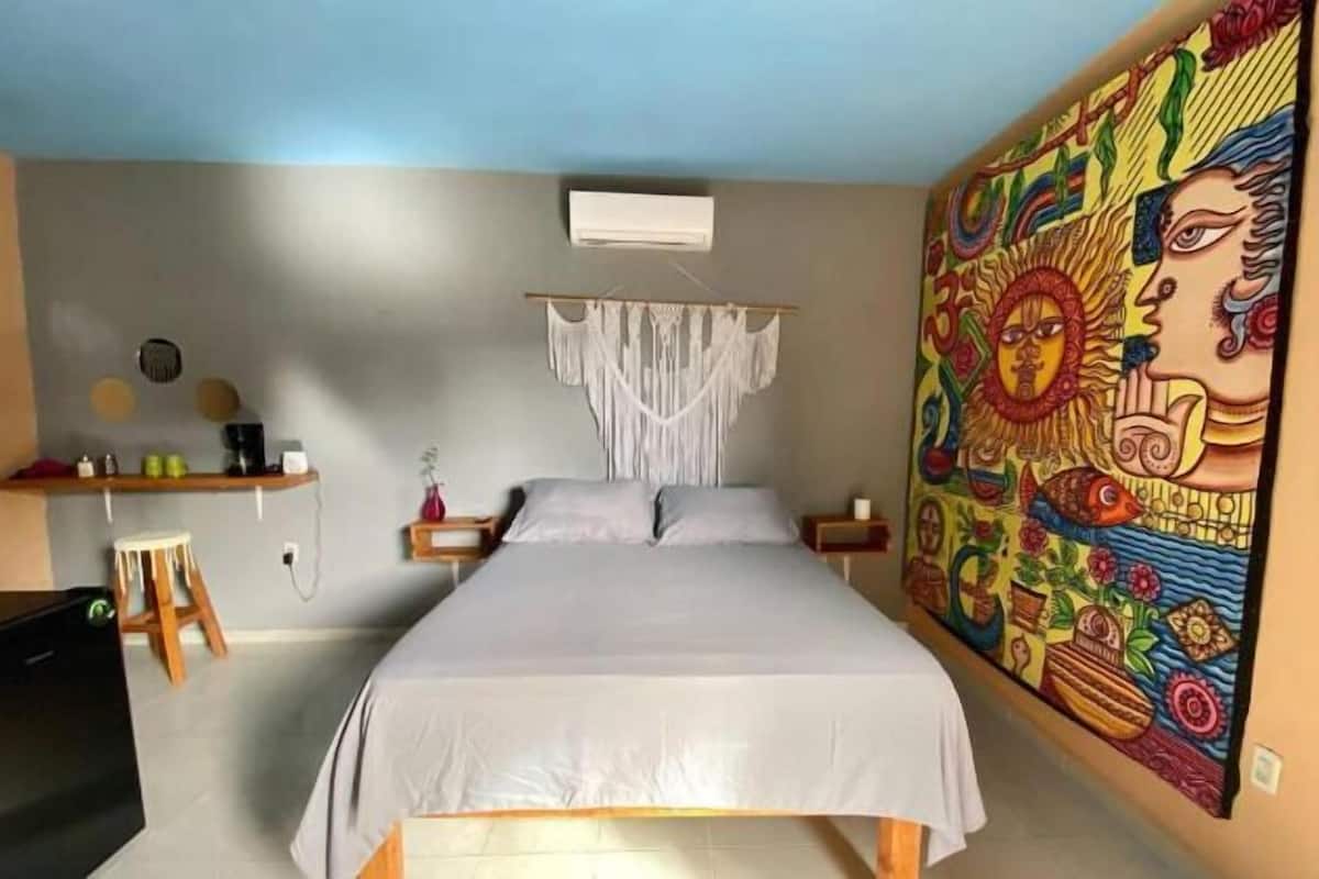 A bedroom with a bed, a macrame wall hanging, a colorful mural, and a small stool.