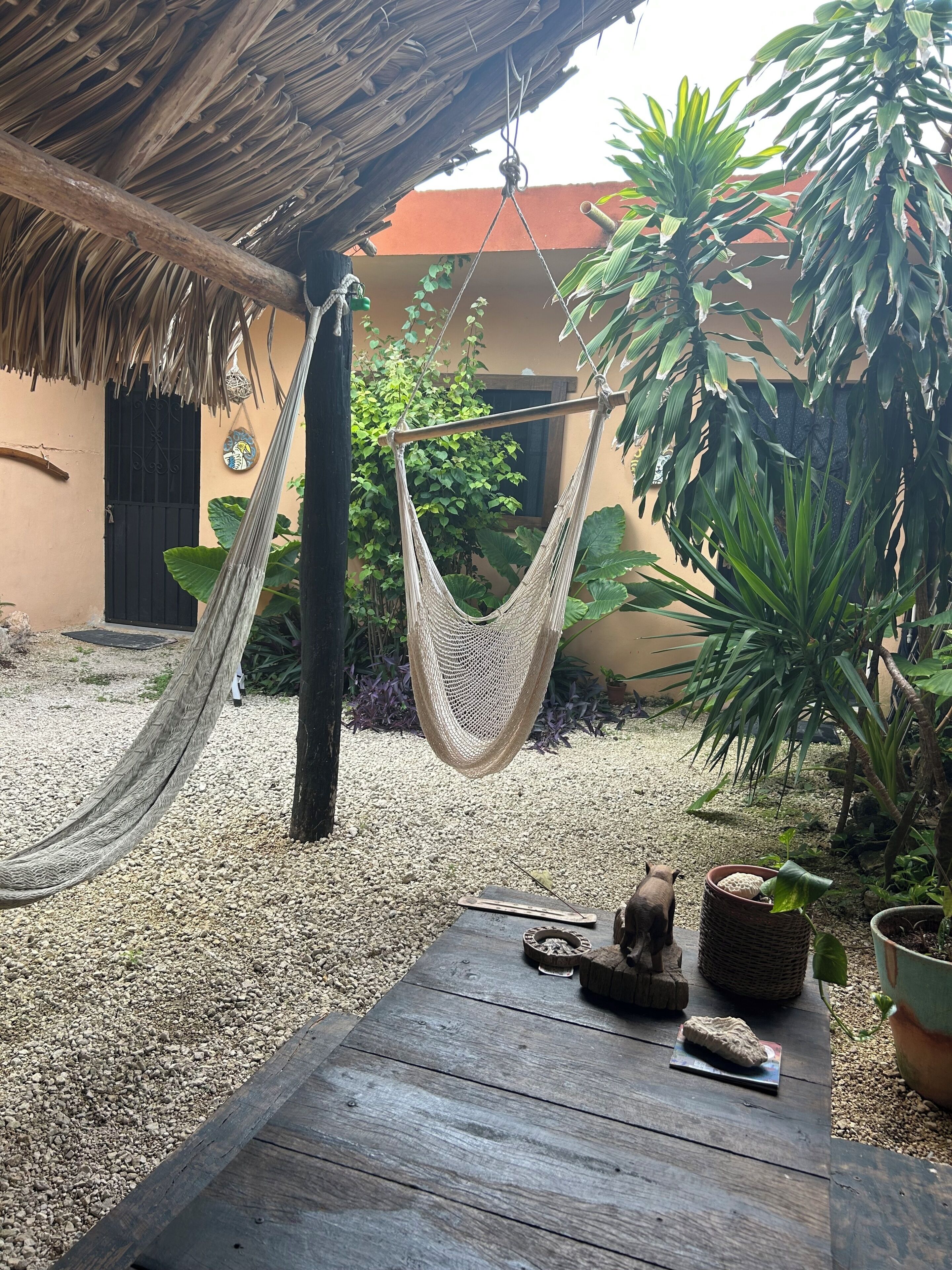 Un hamac suspendu sous un toit de chaume, une table en bois avec un rocher, et une plante en pot.