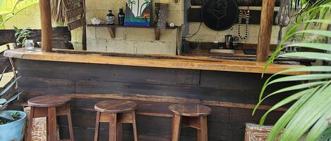 A rustic outdoor bar area with a thatched roof, wooden stools, and a counter with various items.