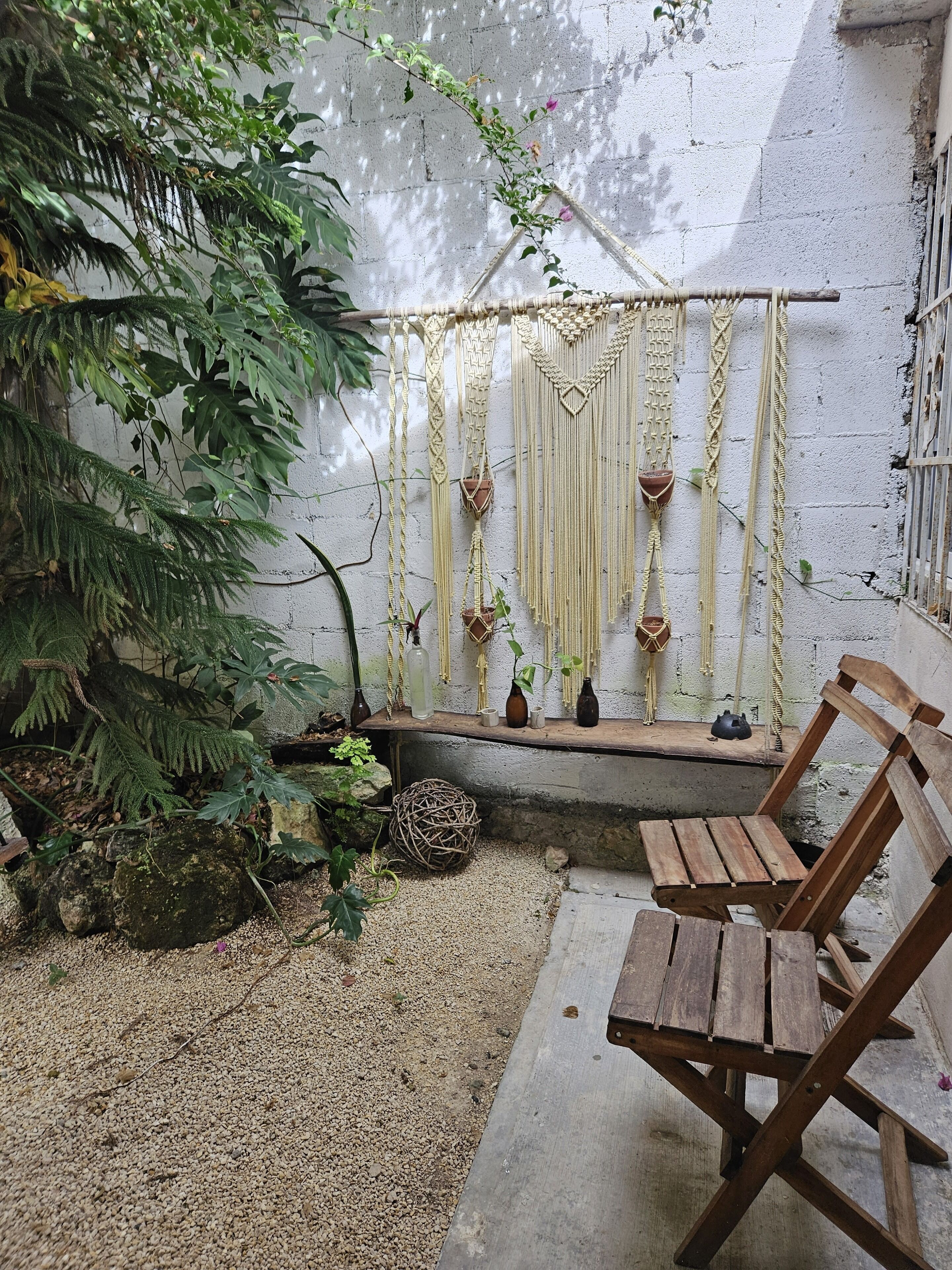 A rustic outdoor seating area with a wooden bench, a woven ball, and a wall adorned with macramé hangings and small potted plants.