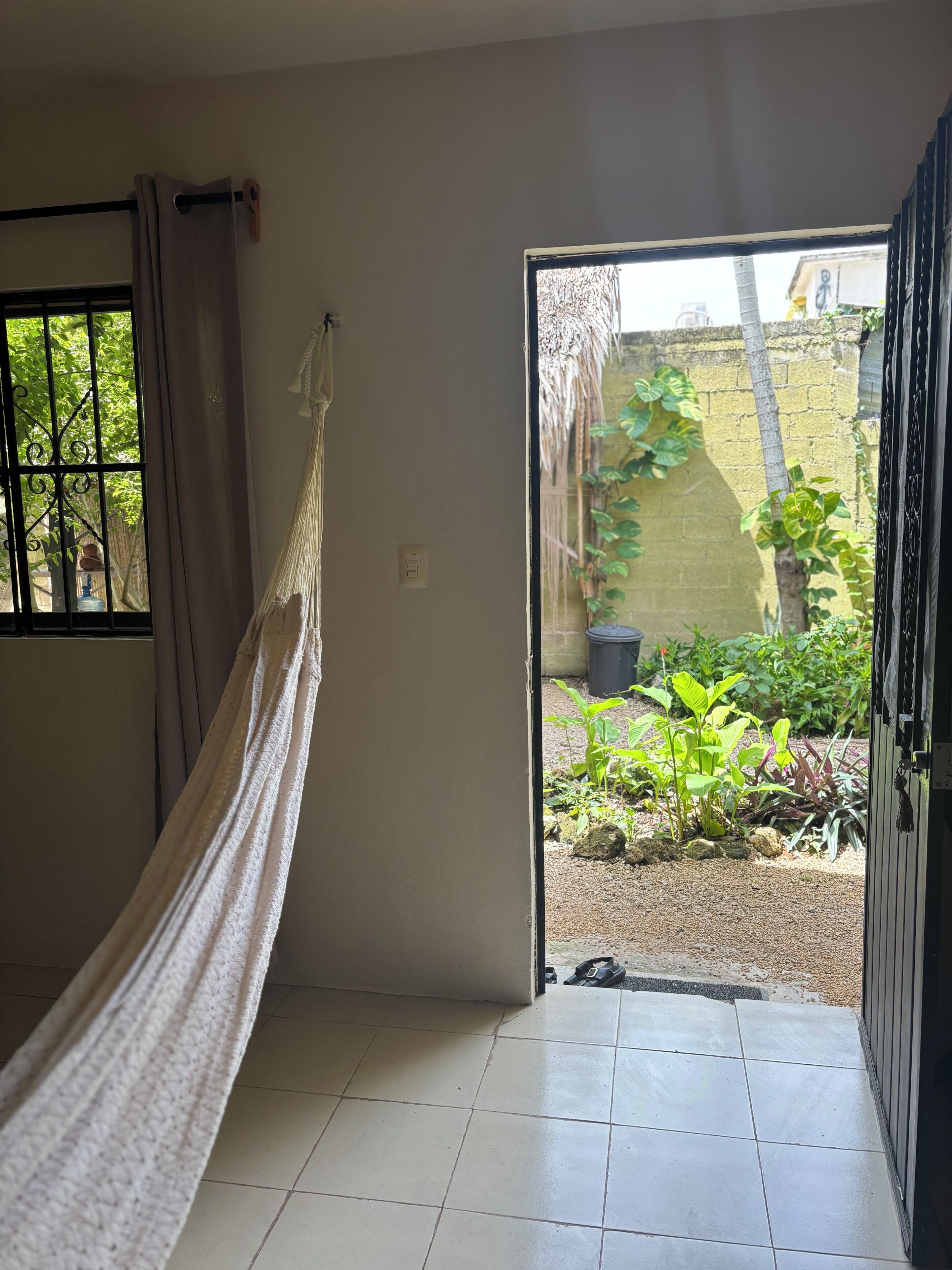 Una habitación con una hamaca, un ventanal con barrotes, una puerta que da a un espacio exterior con plantas y un muro de ladrillo.