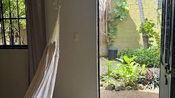 Una habitación con una hamaca, un ventanal con barrotes, una puerta que da a un espacio exterior con plantas y un muro de ladrillo.