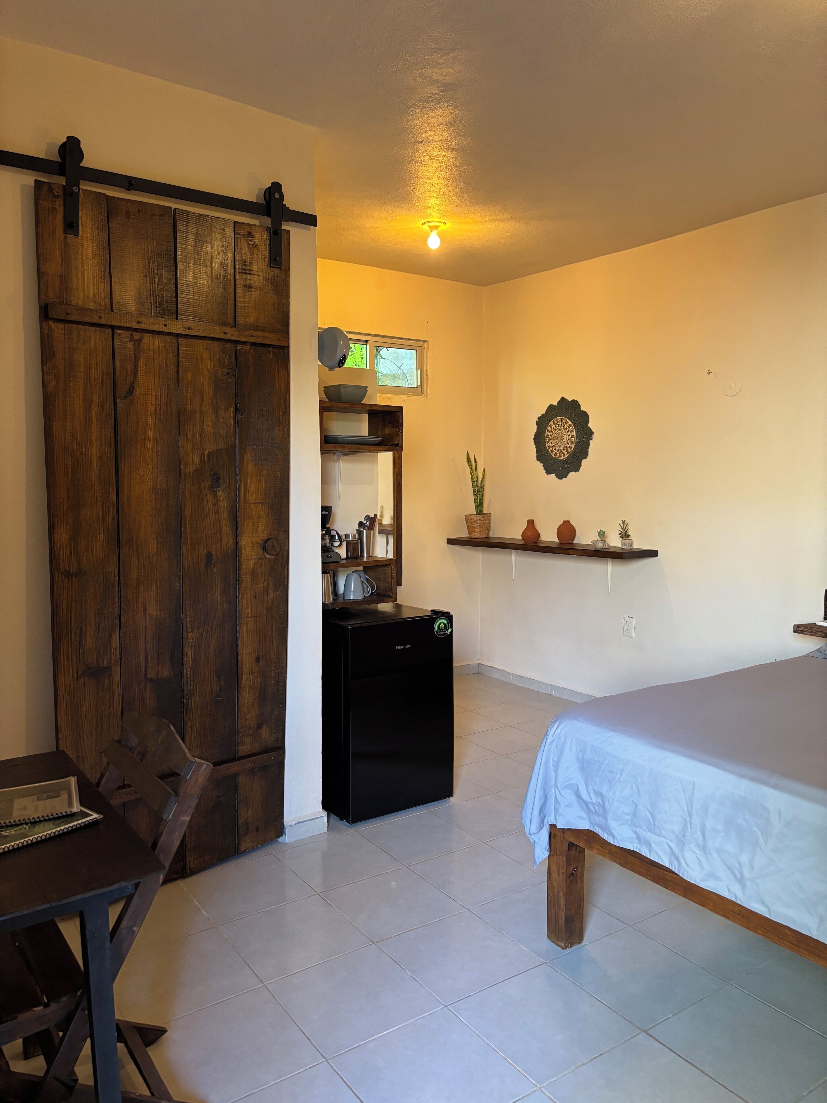 Een slaapkamer met een bed, bureau met stoel, kast, kleine koelkast en een plank met decoratieve voorwerpen.