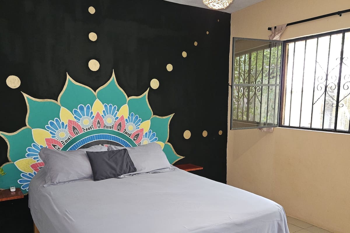 A bed with a light blue bedspread in a room with a black wall and a colorful mural.