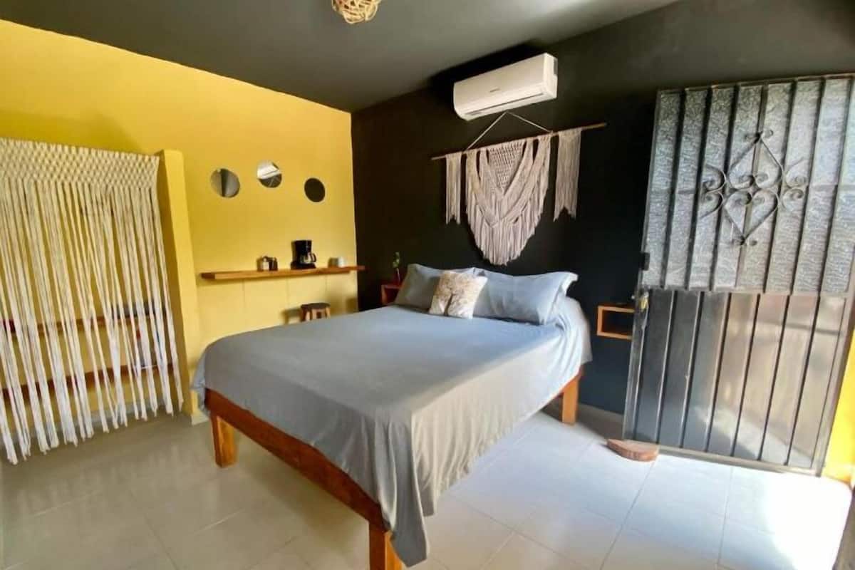 A bedroom with a bed, a small table with a coffee maker, a wall-mounted air conditioner, and a decorative macrame piece.