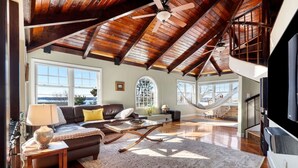 A spacious living room with a high ceiling, wooden beams, a large window, a hammock, and a television. - The widow’s walk your very own spa experience (Marblehead)