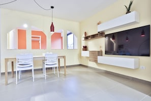 A modern living room with a dining area, a TV mounted on the wall, and a ceiling light fixture.