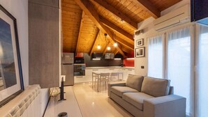 Een modern loft appartement met een hoge plafond, zichtbare houten balken, een keuken met moderne apparatuur, een grijze bank en grote ramen.