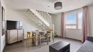 A modern living room with a staircase, a dining area, a flat-screen TV, and a large window with curtains.