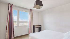 A hotel room with a bed, a desk, a window with curtains, and a view of buildings.