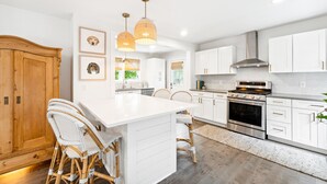 A modern kitchen with white cabinetry, a central island, wicker chairs, pendant lighting, and a stainless steel oven. - Single Family Downtown House with Private Yard (Charleston)