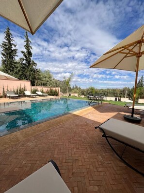 Piscina com espreguiçadeiras e guarda-sóis, cercada por um terraço de tijolos e vegetação.