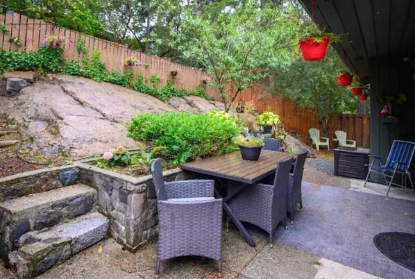 A patio with a wooden table and wicker chairs, surrounded by a stone retaining wall and lush greenery. - Nature’s nook near city (Victoria)
