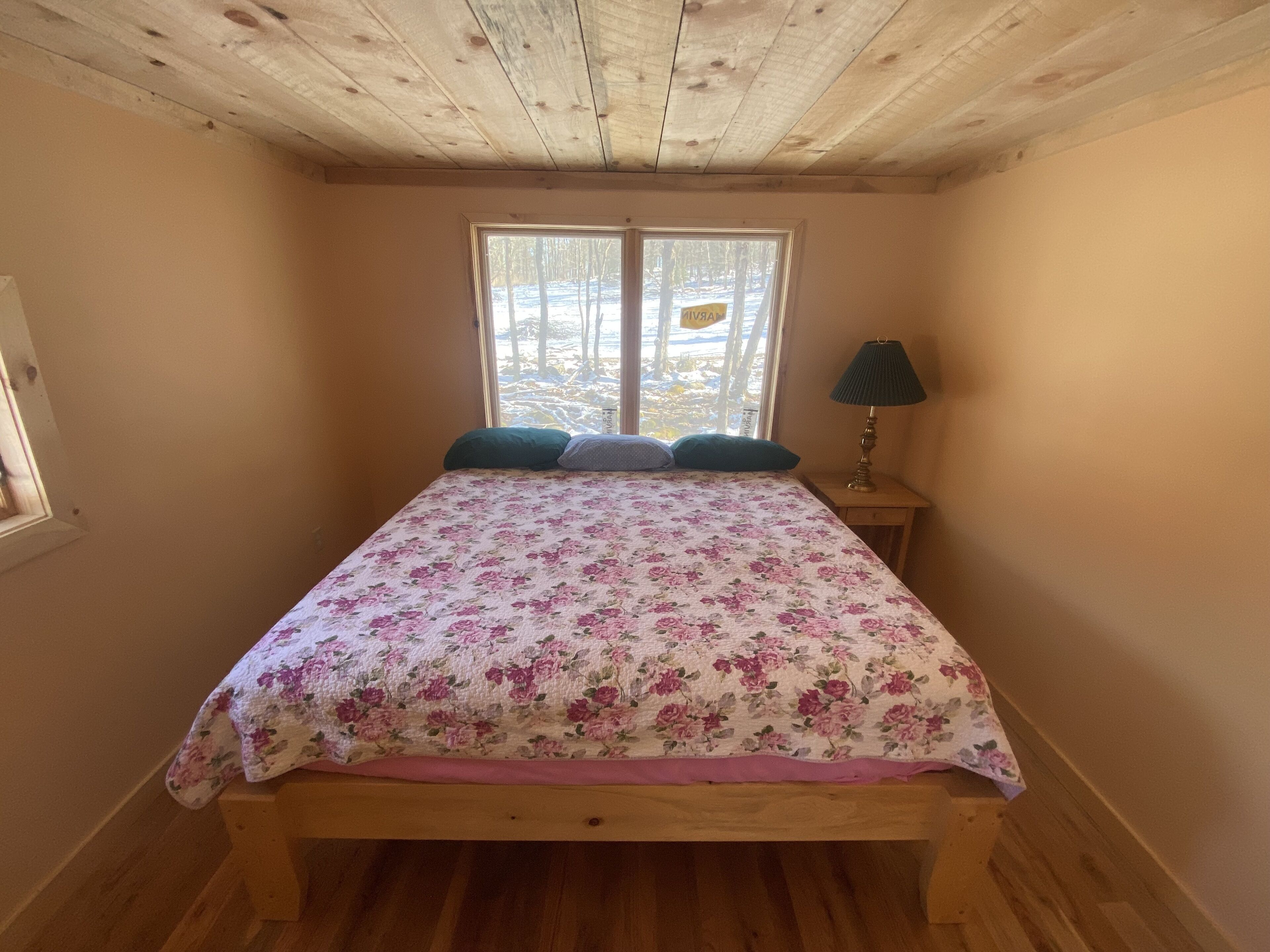 A bedroom with a wooden bed, floral bedding, a nightstand with a lamp, and a window with a view of trees.