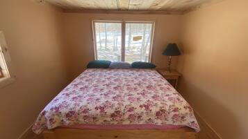 A bedroom with a wooden bed, floral bedding, a nightstand with a lamp, and a window with a view of trees.