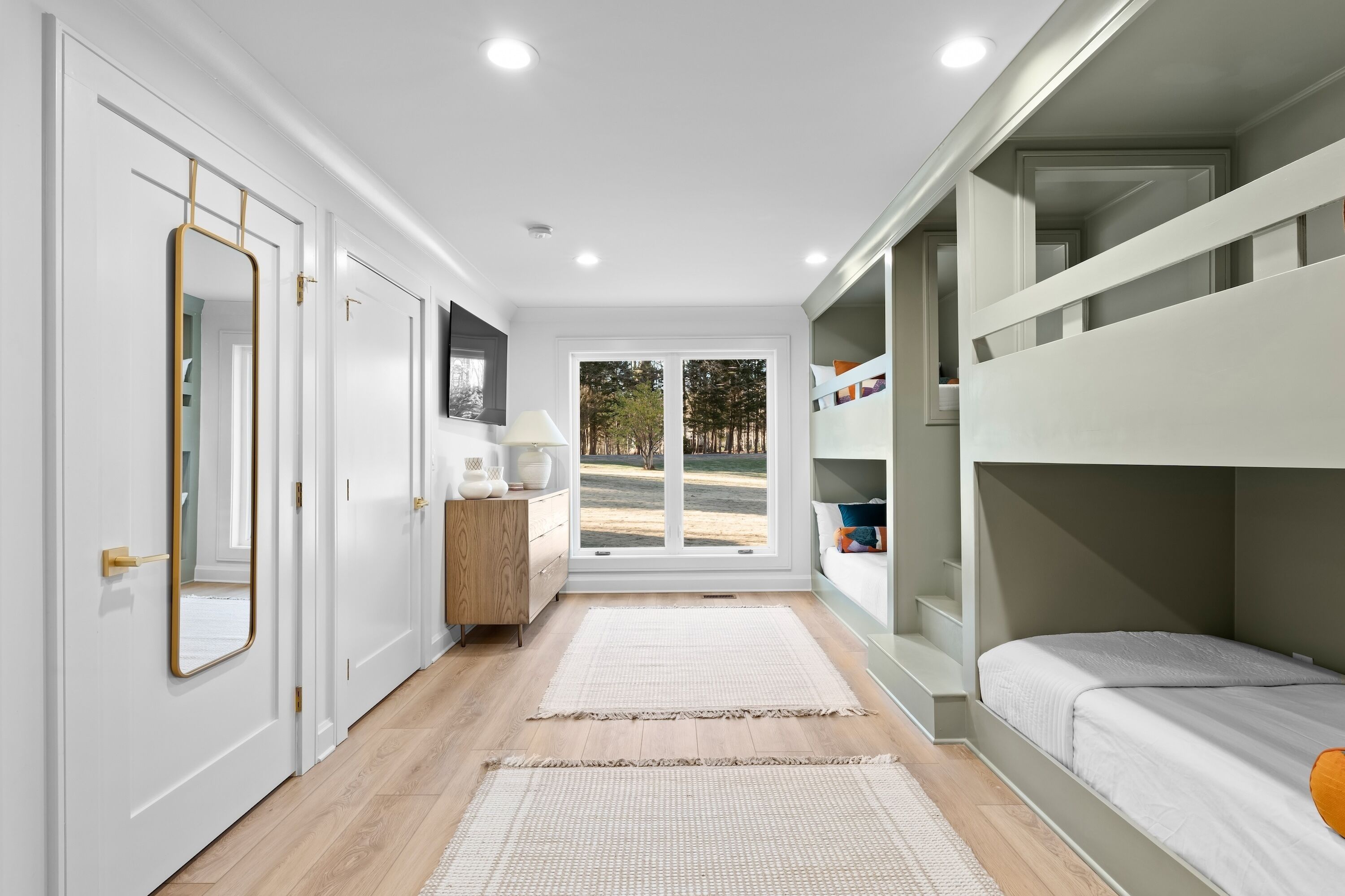 A bedroom with a bunk bed, a dresser, a mirror, and a window with a view of trees.