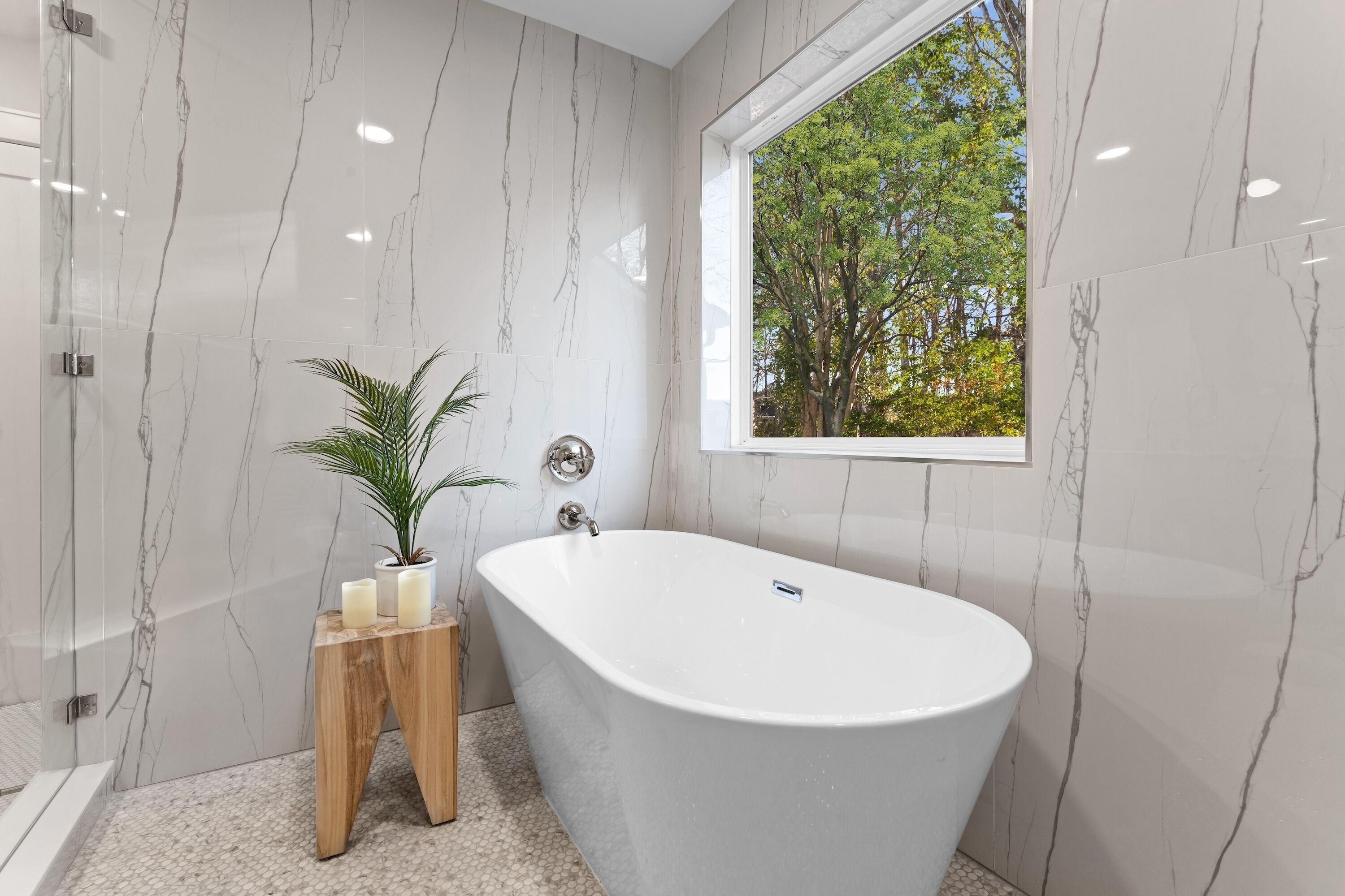 Un baño moderno con una bañera independiente, paredes de mármol, una mesita de madera con velas y una ventana con vistas a árboles.