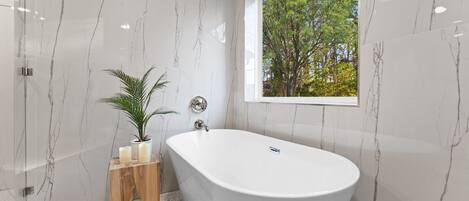 Un baño moderno con una bañera independiente, paredes de mármol, una mesita de madera con velas y una ventana con vistas a árboles.