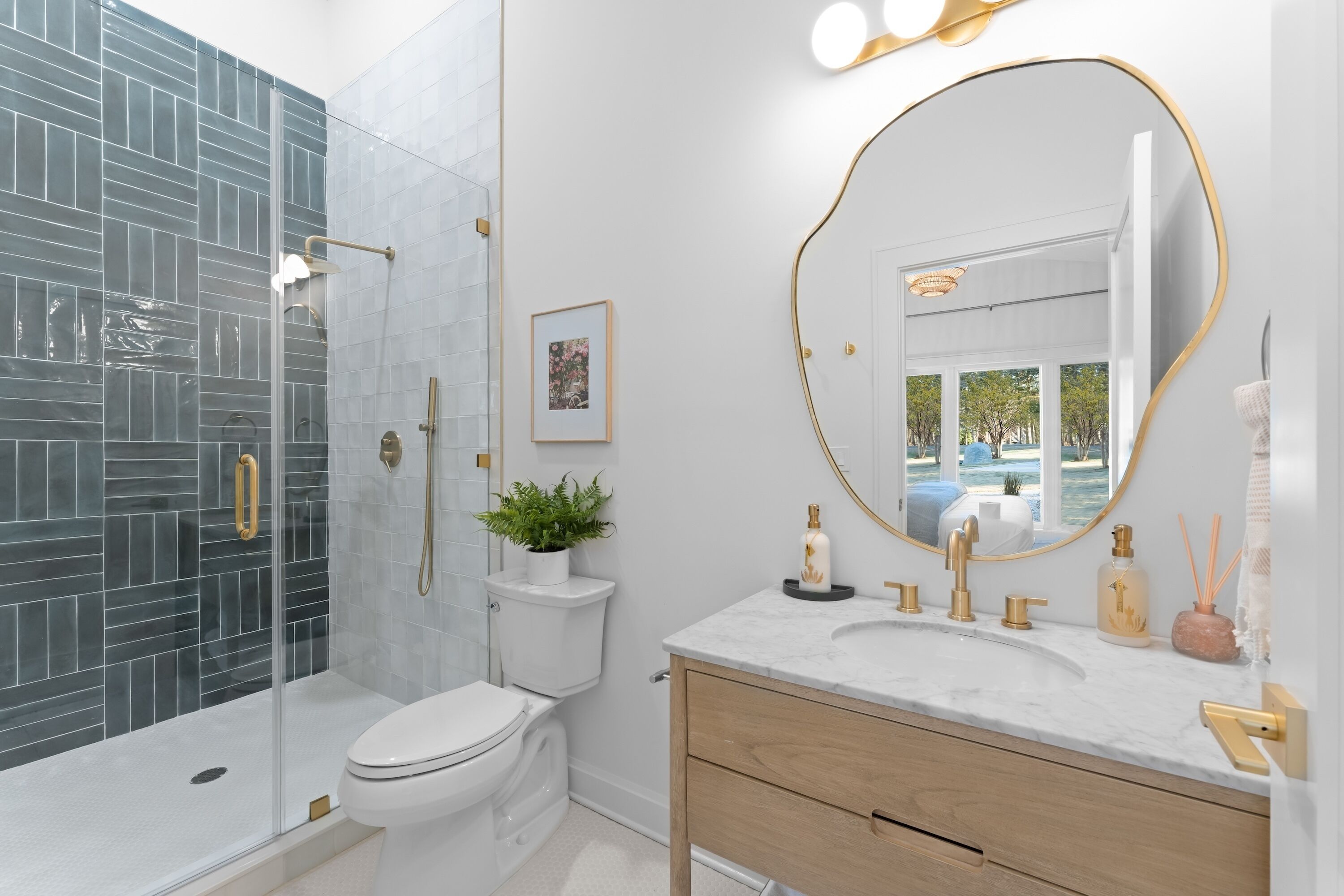 A modern bathroom with a glass shower enclosure, a white toilet, a vanity with a gold-framed mirror, and a potted plant.