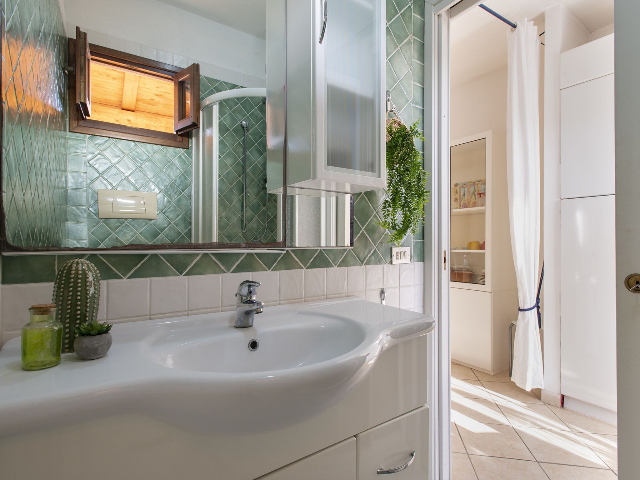 A bathroom with a white sink, green mosaic tiles, a cactus, and a mirror.