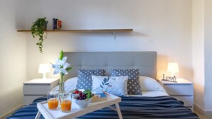 A neatly made bed with a tray of orange juice, a bowl of fruit, and a white flower. There are two bedside lamps, a shelf with books and a plant, and a gray polka dot pillow. - CSB - 9 - Baia de Bahas by Interhome (Golfo di Marinella)