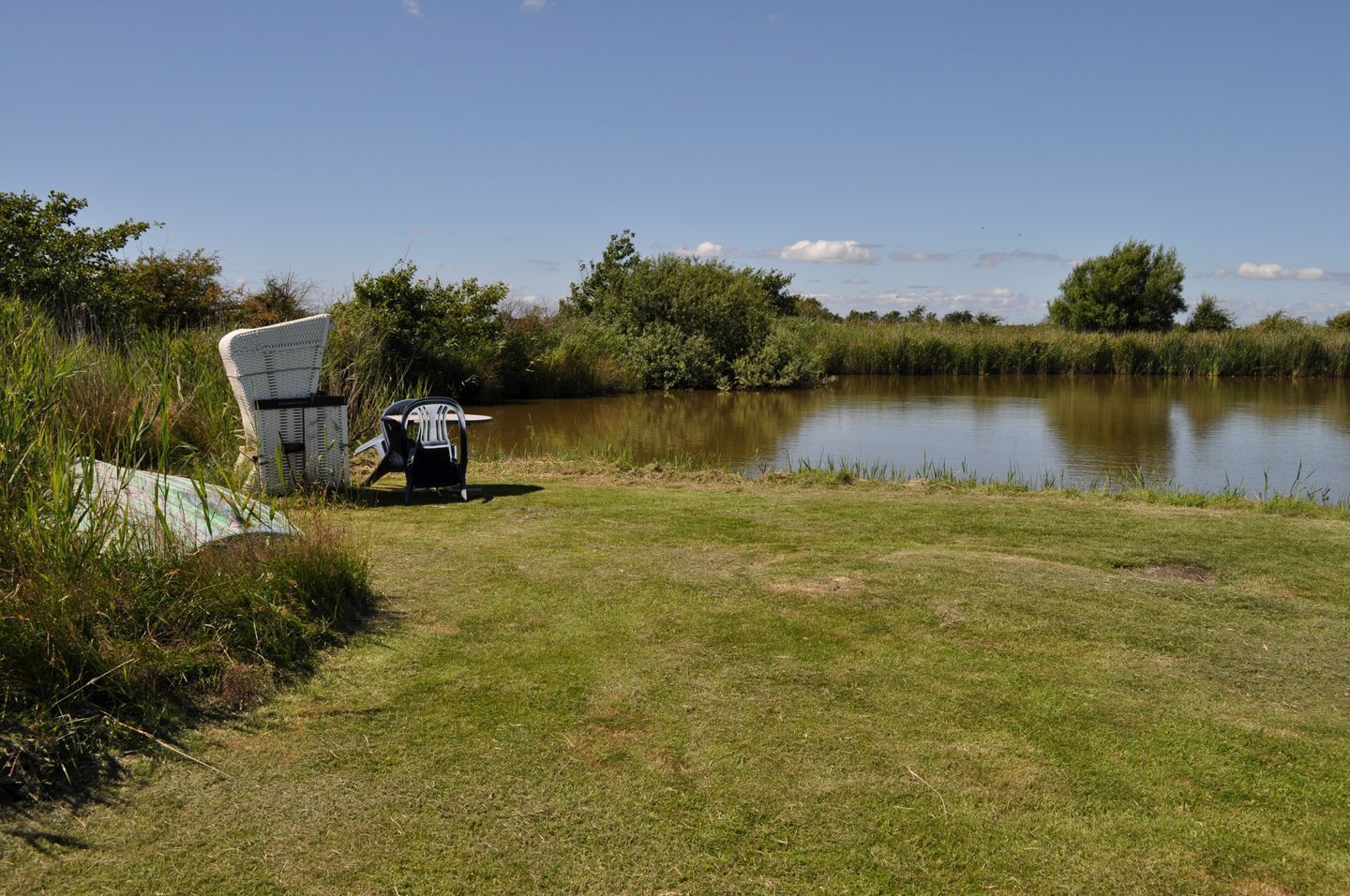 A grassy area with a white chair and a black chair near a calm body of water, surrounded by greenery.