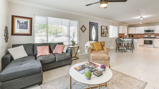 A modern living room with a grey sectional sofa, a beige armchair, and a coffee table. There is a ceiling fan, a dining area with a kitchen in the background, and a large window with blinds. - 5 Mi to Dtwn: Pet-Friendly Phoenix Gem w/ Patio (Phoenix)