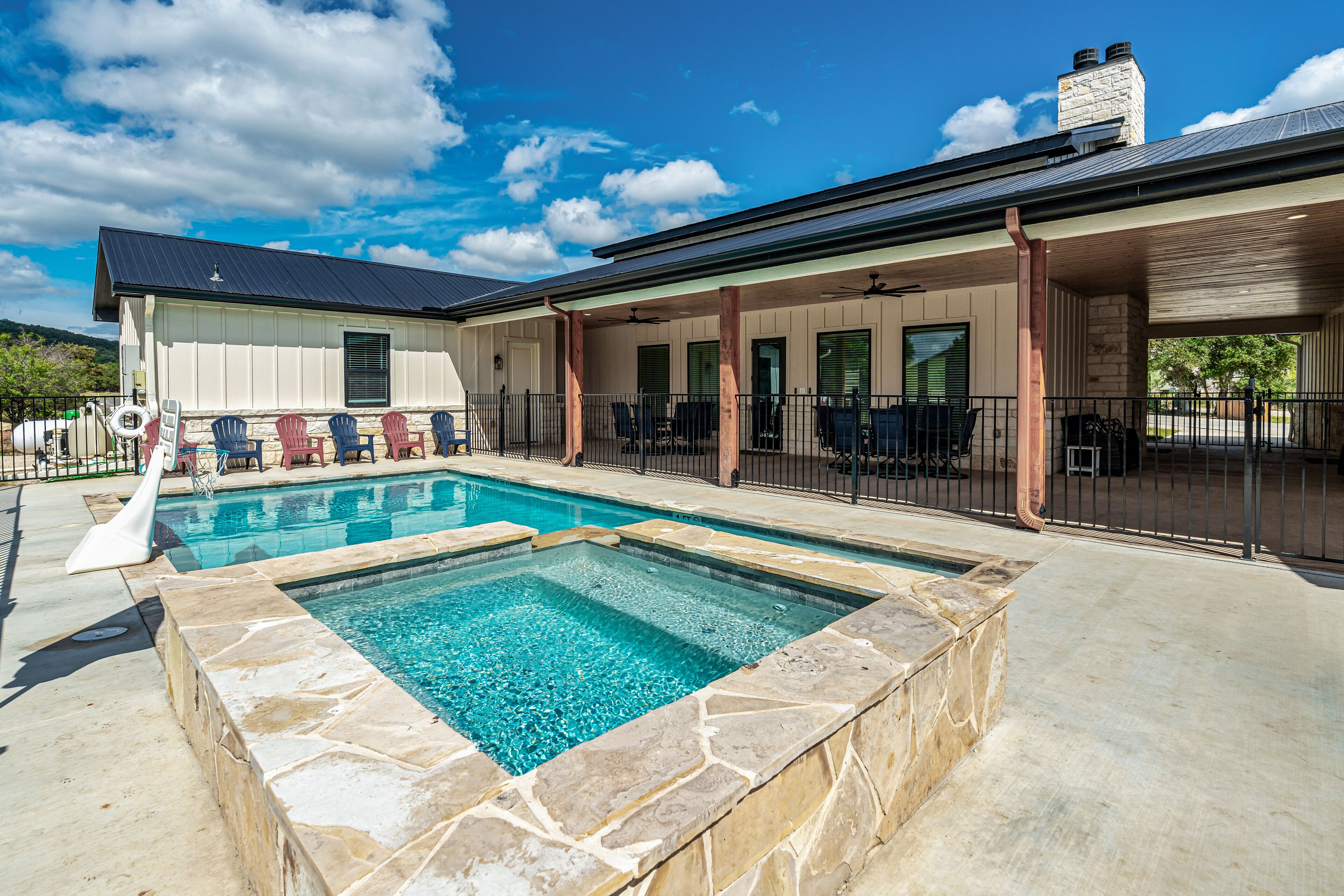 A swimming pool with stone tiles, a covered patio with chairs, and a house with a stone chimney.