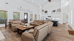 A spacious living room with a large beige sectional sofa, a wooden coffee table, and a dining area with a table and chairs. The kitchen is visible in the background with white cabinets and a large island. - Hot tub, pool, Sonos speaker system (Concan)
