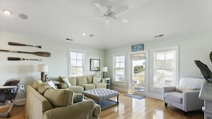 A spacious living room with a beige sofa, a striped armchair, and a wooden floor. There are two windows with blinds, a ceiling fan, and a view of the outdoors.