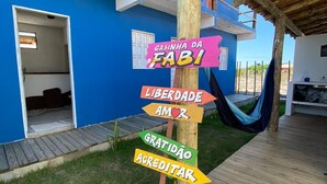 Um painel colorido com frases como "CASINHA DA FABI" e "LIBERDADE AMOR" em frente a um edifício azul com varanda de madeira e uma rede.