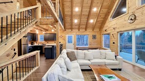 Ruime cabin inrichting met een houten trap, een zithoek met bank en salontafel, een keuken, en grote ramen met uitzicht op het omliggende bos.