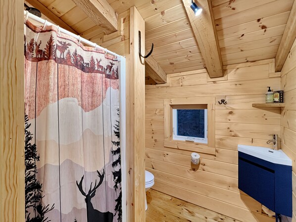 Een houten badkamer in een chalet, met een douchegordijn met een boslandschap, een klein raam, een toilet en een wastafel met kastje.