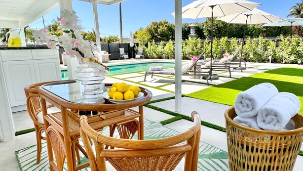 A patio area with a table, chairs, a basket of towels, and a pool in the background. - MIAMI LUX HOUSE / Pool / Lounge / Bar (Miami)
