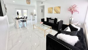 A modern living room with a black sofa, a glass coffee table, and a dining area with a white table and chairs. - MIAMI LUX HOUSE / Pool / Lounge / Bar (Miami)