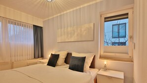 Une chambre à coucher comprenant un lit, des tables de chevet, une lampe, une fenêtre avec des stores et une vue sur l’extérieur.