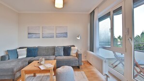 Un salon moderne avec un canapé d’angle gris, une table basse en bois et un pouf gris. Trois œuvres d’art encadrées ornent le mur, ainsi qu’une lampe sur pied et une grande fenêtre donnant sur l’extérieur.