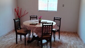 A dining area with a round table, four chairs, a vase with flowers, and a window with blinds. - Recanto Silvia Timbó (Tapera)