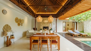 A spacious indoor area with a wooden ceiling, a dining table with chairs, a sofa, and a pool area with lounge chairs.