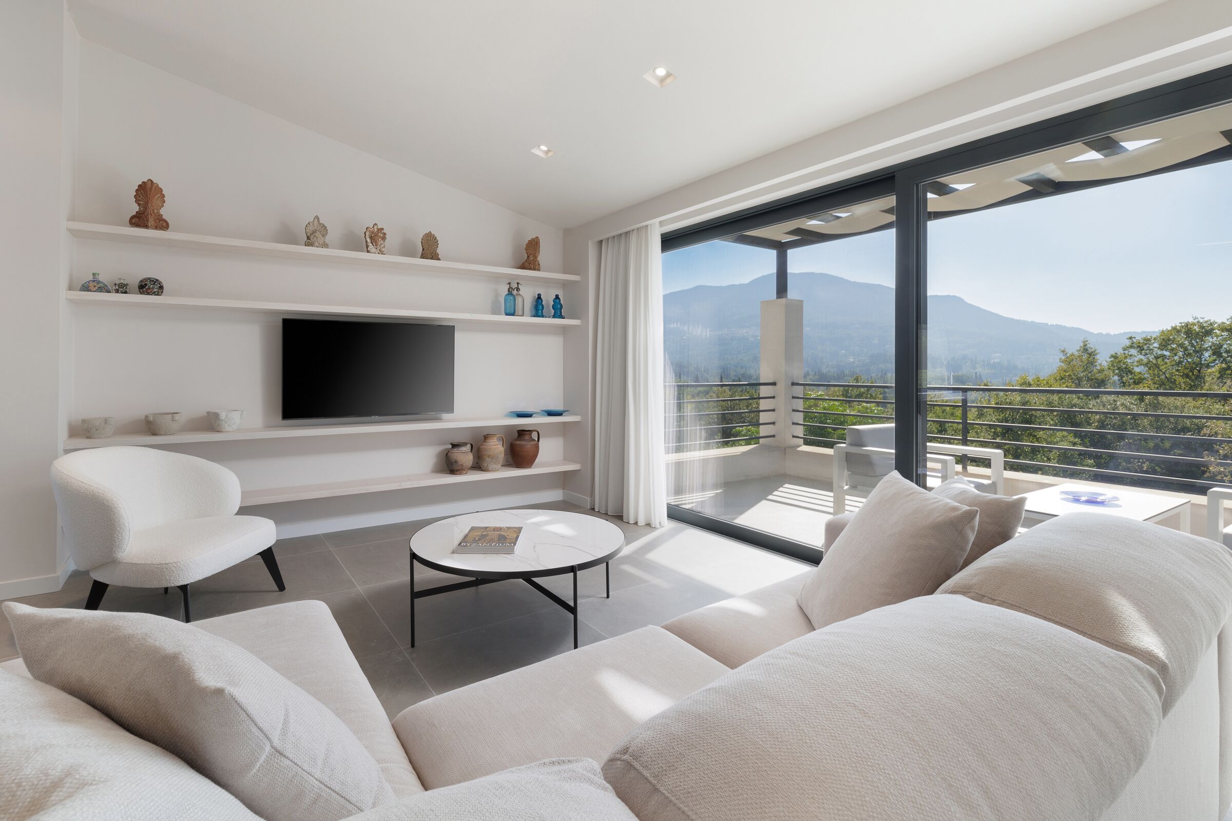Un salón moderno con televisor de pantalla plana, mobiliario blanco y un balcón con vistas a la montaña.