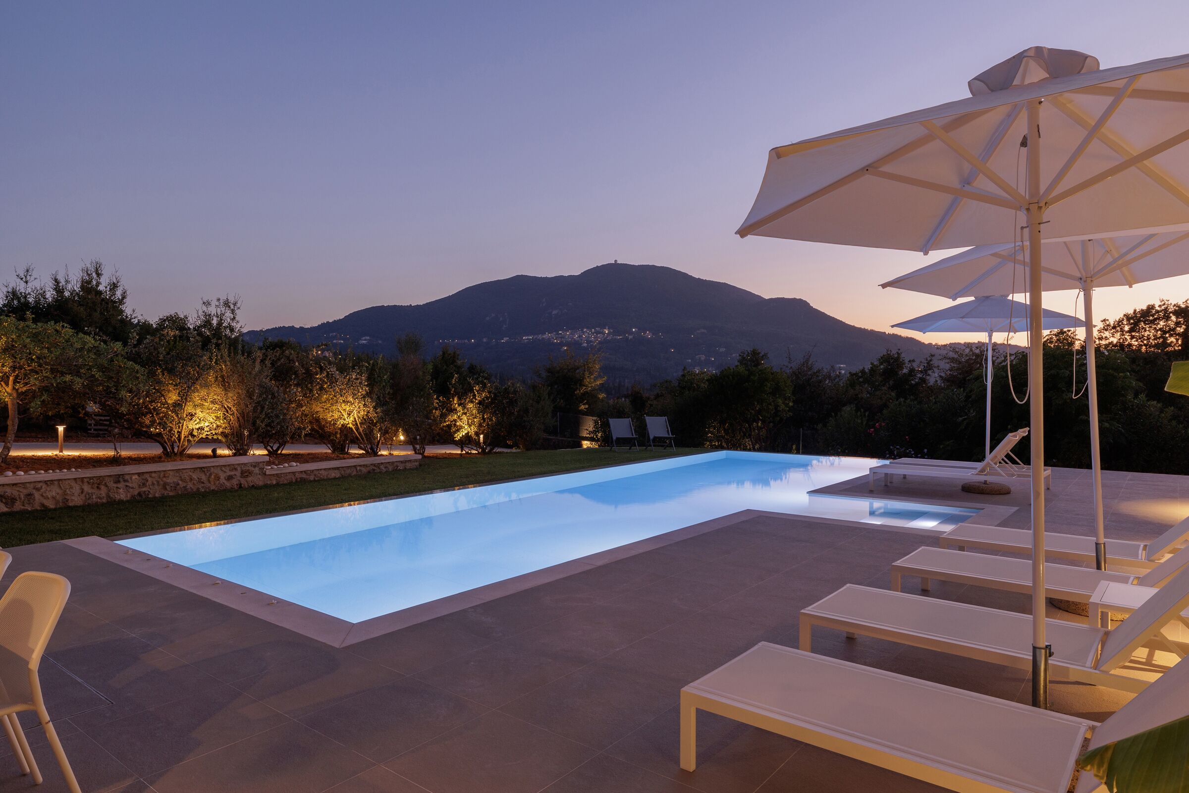 A swimming pool with lounge chairs and umbrellas, surrounded by landscaped gardens and a mountain in the background.