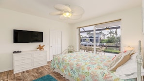 Une chambre avec un lit, une télévision, un ventilateur de plafond, une fenêtre donnant sur une maison et des palmiers, et une commode blanche.