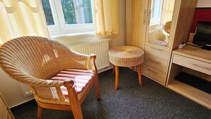 Ein Zimmer mit einem Korbstuhl, einem kleinen runden Tisch, einem Holzbüro mit einem Computer und einem Fenster mit Vorhängen.