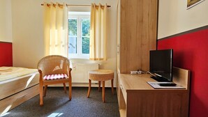 Ein Zimmer mit einem Bett, einem Korbsessel, einem kleinen Tisch mit Fernseher und einem Fenster mit Vorhängen.