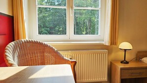 Una habitación con una silla de mimbre, una mesita de noche con lámpara, un radiador y una ventana con vista a árboles.