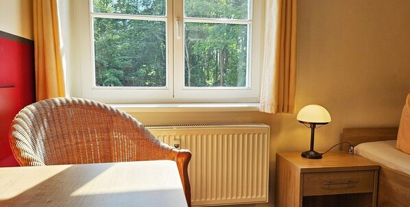 A room with a wicker chair, a bedside table with a lamp, a radiator, and a window with a view of trees.