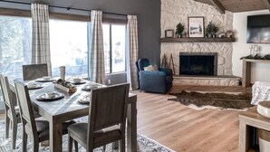 Un área de comedor con una mesa y sillas de madera, una chimenea con repisa de piedra y un ventanal con cortinas.