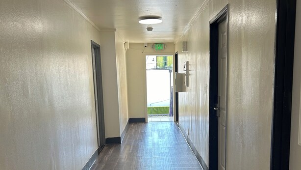 A long, narrow hallway with a wooden floor and beige walls. There are doors on both sides, and an exit sign is visible on the wall. - Budget Inn Hollywood (Los Angeles)