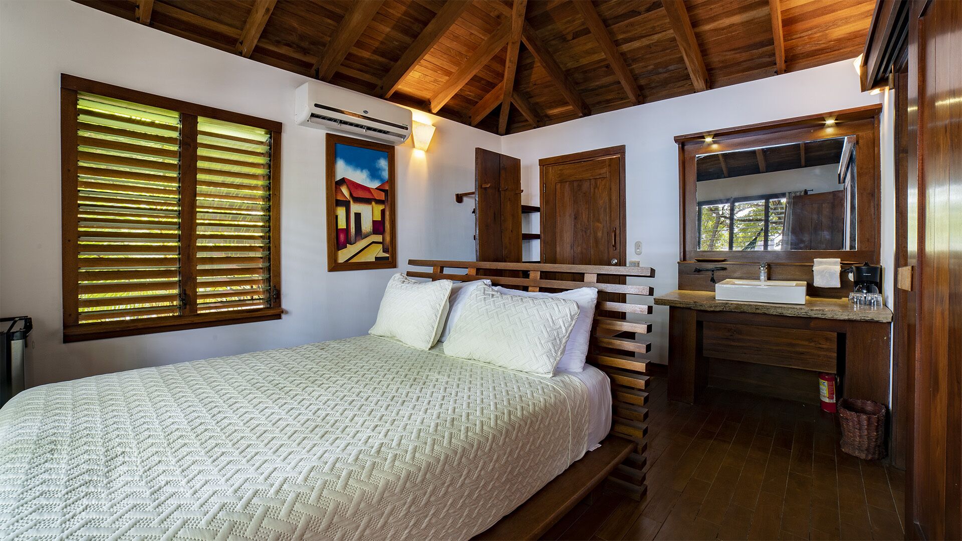 Un dormitorio con una cama de madera, un ventanal con persianas, un equipo de aire acondicionado, un cuadro y una cómoda de madera con espejo.